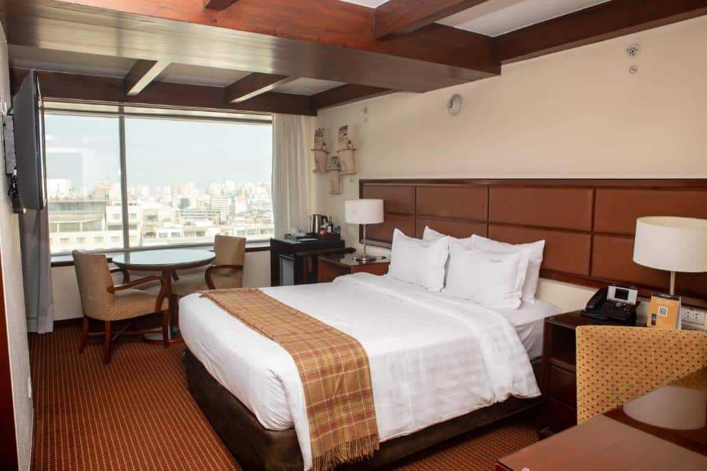 Um quarto de hotel com uma cama de casal, cadeiras, escrivaninha, janela com vista para a cidade e vigas de madeira no teto.