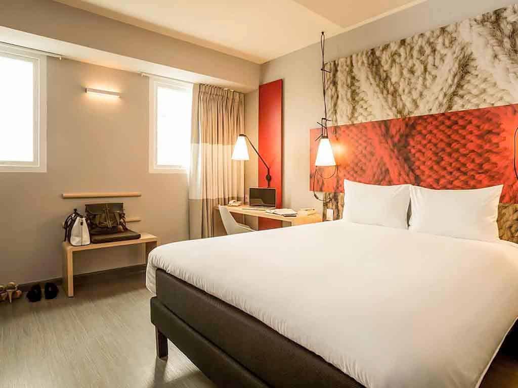 Quarto do Ibis Lima Reducto, onde ficar em Lima. Quarto de hotel moderno com uma cama grande, escrivaninha, duas janelas, arte na parede e itens pessoais bem organizados em um banco.