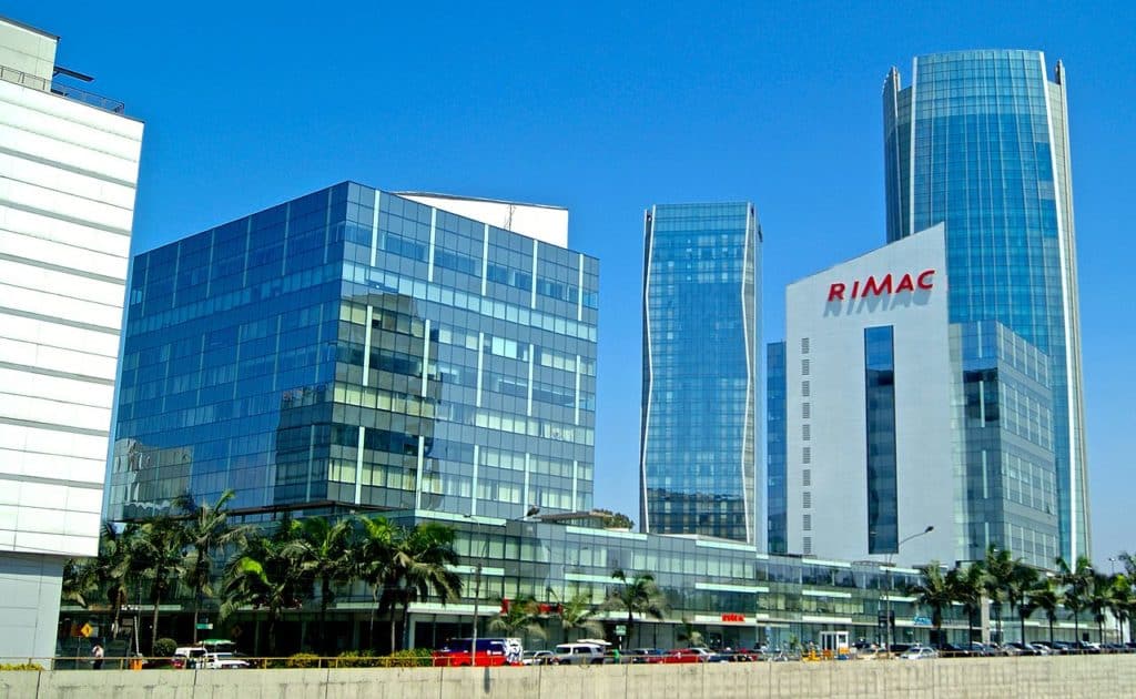 Prédios comerciais espelhados no bairro de San Isidro, em Lima. Modernos prédios de escritórios de vidro sob um céu azul claro, com a placa "RIMAC" em uma torre alta e palmeiras na frente.