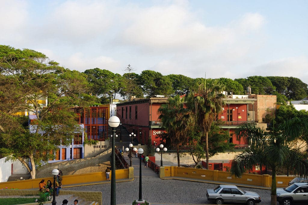 Foto de área do bairro Barranco, em Lima. As pessoas caminham perto de edifícios coloridos, árvores e carros estacionados em uma rua de paralelepípedos sob um céu parcialmente nublado.