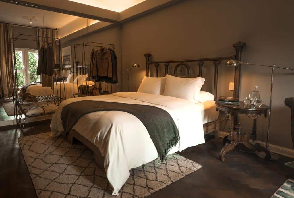 Um quarto elegante com uma cama grande, tapete estampado, cabideiro de metal e uma mesa lateral redonda de madeira com copos.