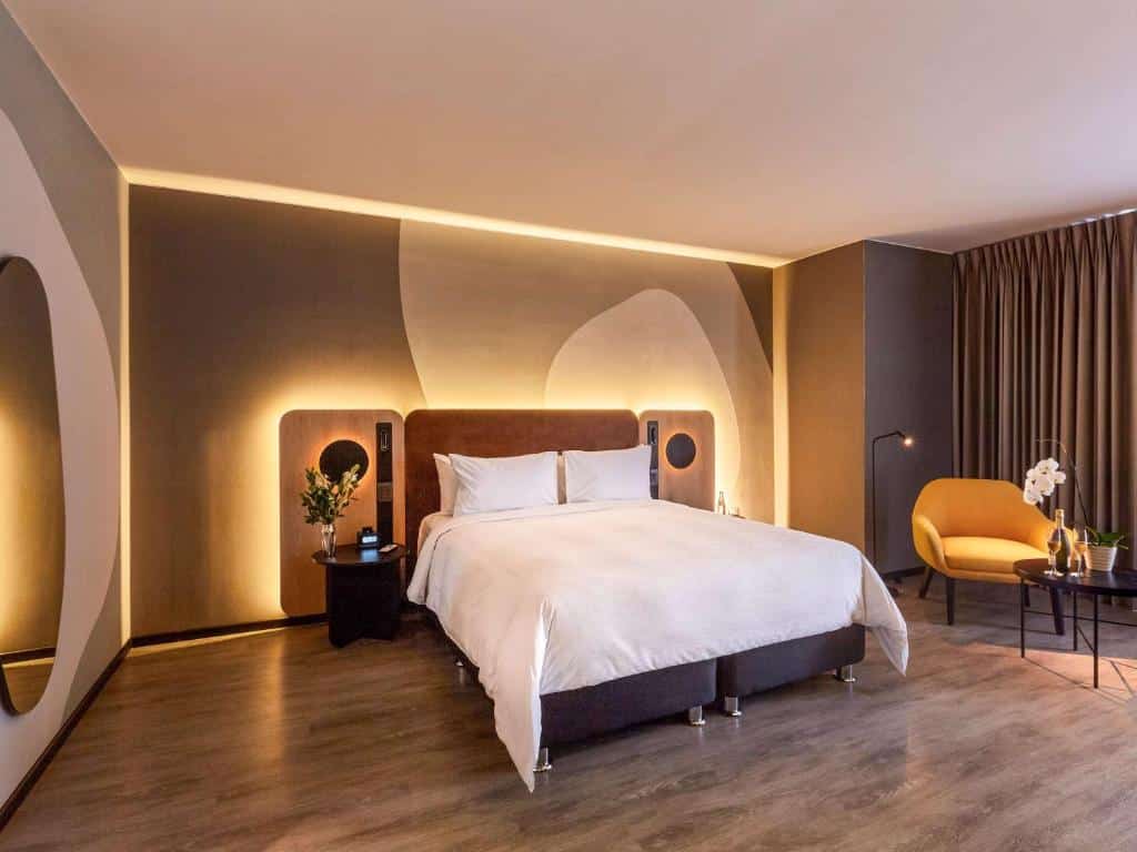 Quarto de hotel moderno com uma cama king-size, iluminação de destaque, cadeira amarela, mesa redonda e piso de madeira.