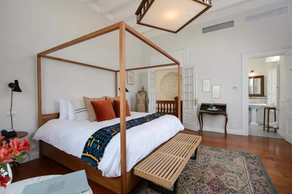 Um quarto com cama de madeira com dossel, banco, tapete, escrivaninha e vista para um banheiro adjacente.
