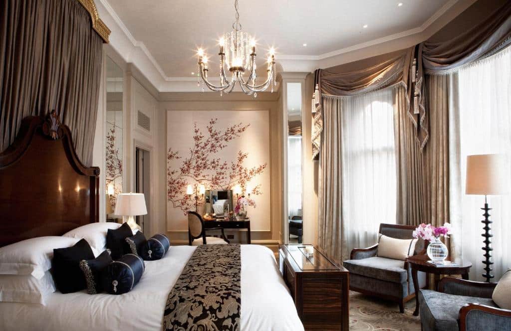 Quarto luxuoso do The Langham London com janelas com cortinas, um lustre, uma cama de casal com muitas almofadas e travesseiros, uma penteadeira, duas poltronas e um abajur de chão, para representar onde ficar em Londres.