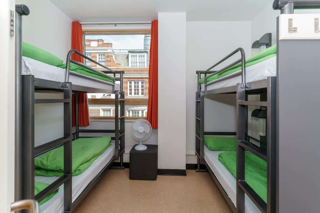 Quarto compartilhado do YHA London Central com duas beliches, uma janela com cortinas um ventilador de chão.