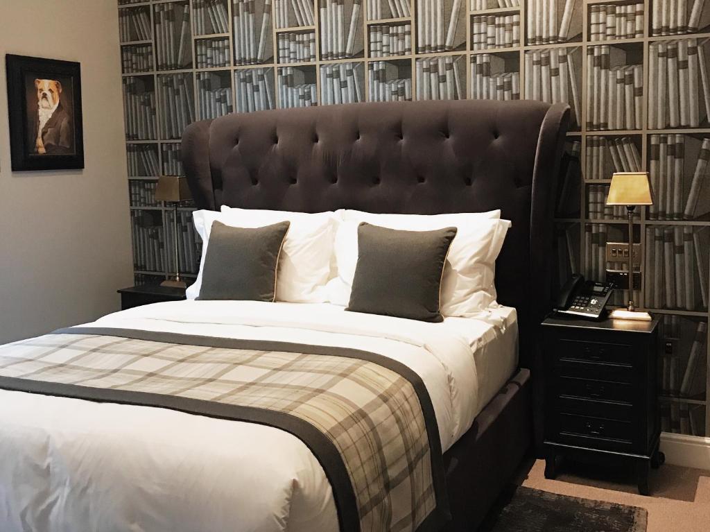 Uma cama bem arrumada, com travesseiros cinza e estampa xadrez, ladeada por duas mesinhas de cabeceira pretas e um papel de parede com estampa de livros, no The Grafton Arms Pub & Rooms, para representar onde ficar em Londres.