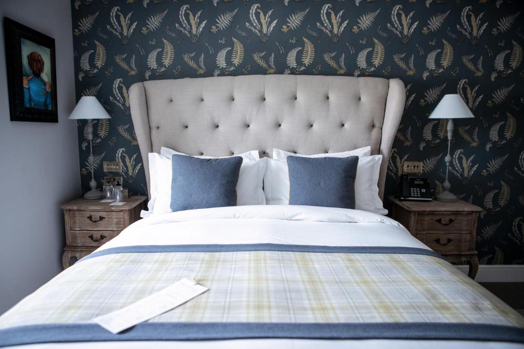 Uma cama bem arrumada com roupa de cama azul e branca fica entre duas mesas de cabeceira, contra uma parede com papel de parede estampado, no The Hayden Pub & Rooms, para representar onde ficar em Londres.