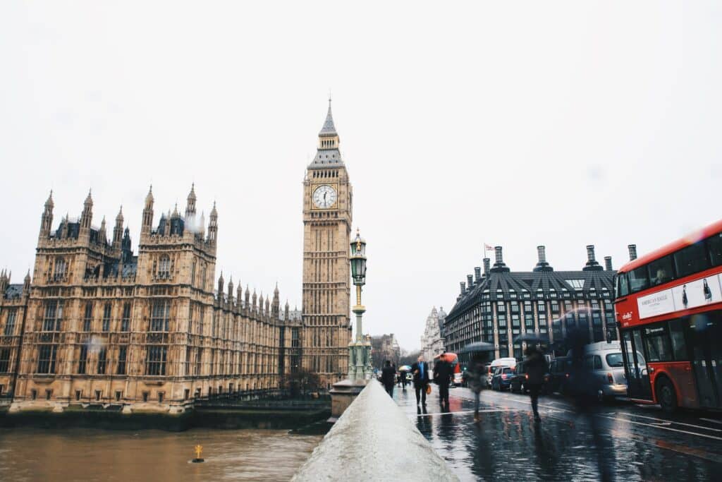 Prédio histórico que conta com uma torra de relógio alto, o Big Ben, de frente para o rio Tâmisa e com uma ponte movimentada, com ônibus, carros e pessoas passando.