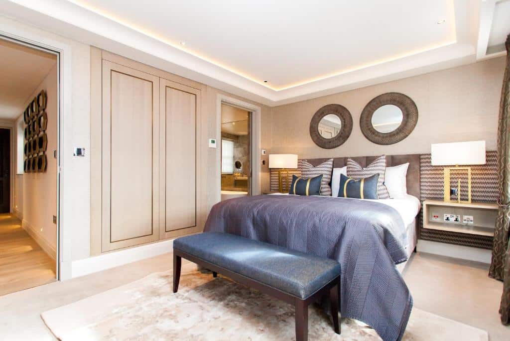 Quarto de luxo do Hope House Residences by Q Apartments com uma cama de casa, dois espelhos redondos, duas mesinhas de cabeceira com abajures, um pequeno armário, um tapete e um sofázinho, para representar onde ficar em Londres.