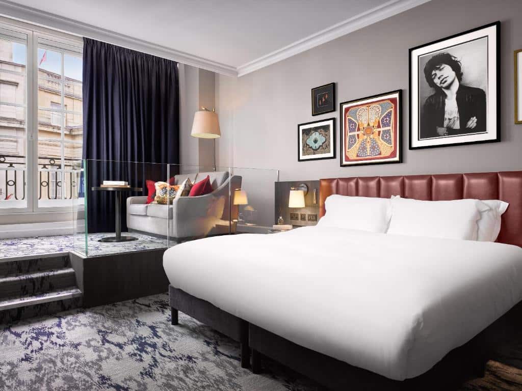 Quarto do The Trafalgar St. James, London Curio collection by Hilton com uma janela ampla com uma sacada com cortinas, um sofá com almofadas, com um abajur, há carpete por todo o ambiente, uma cama de casal com uma cabeceira de couro e alguns quadros na parede, para representar onde ficar em Londres.