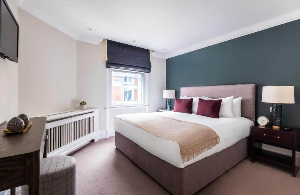 Quarto do Native Mayfair com uma cama de casal, uma janela com persiana, duas mesinhas de cabeceira, um móvel de madeira com um bufê e uma televisão, para representar onde ficar em Londres.