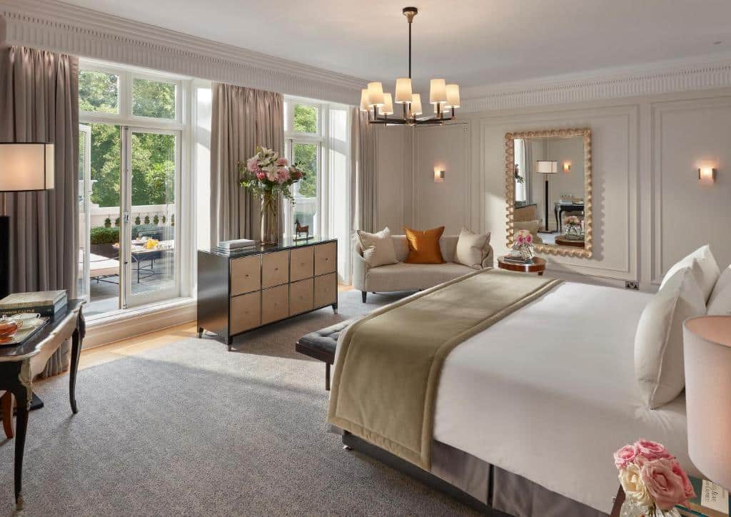 Quarto espaçoso do Mandarin Oriental Hyde Park com uma sacada ampla com vista para árvores, e algumas poltronas ao ar livre, do lado de dentro há uma cômoda, cama de casal, um sofá com almofadas, além de um lustre e um espelho.