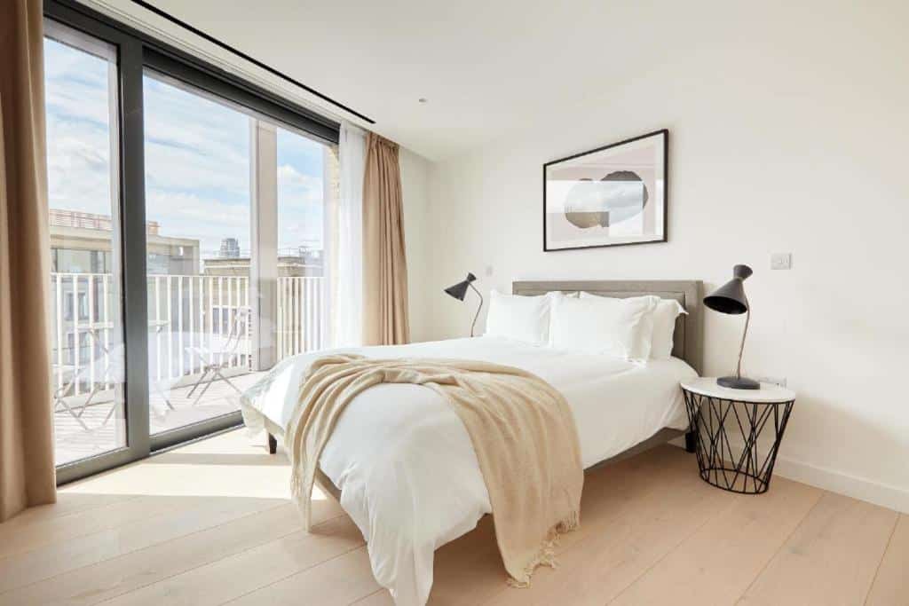 Quarto do Southwark by Q Apartments com uma sacada com cadeiras, uma cama de casal, duas cabeceiras orgânicas com luminárias, um quadro e o chão é de madeira, para representar onde ficar em Londres.