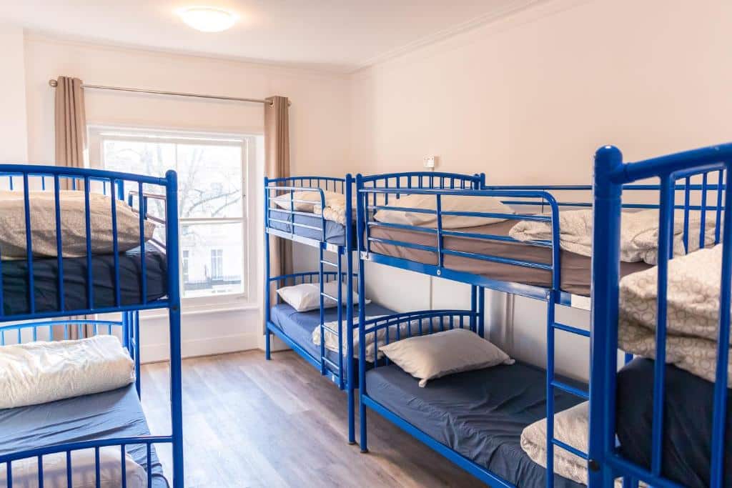 Quarto no Urbany Hostel London com várias beliches azuis com travesseiros, além de uma ampla janela com cortina e o chão é de madeira, para representar onde ficar em Londres.