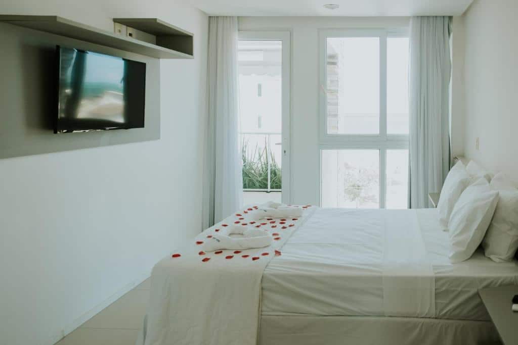 Quarto minimalista no Wave Suítes Hotel na Praia de Ipioca, com uma cama decorada com pétalas de rosa, TV na parede e janelas grandes com cortinas transparentes, para representar onde ficar em Maceió.
