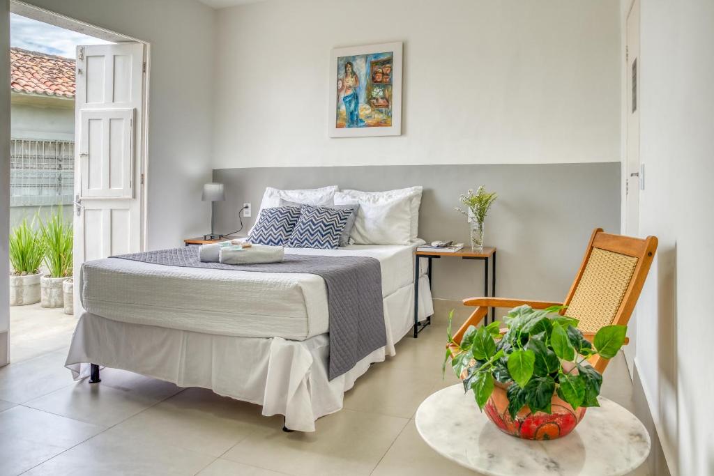 Um quarto bem iluminado com uma cama de casal, cadeira de madeira, vaso de plantas em uma mesa redonda e uma pintura acima da cama, na Pousada Praia Pajuçara.