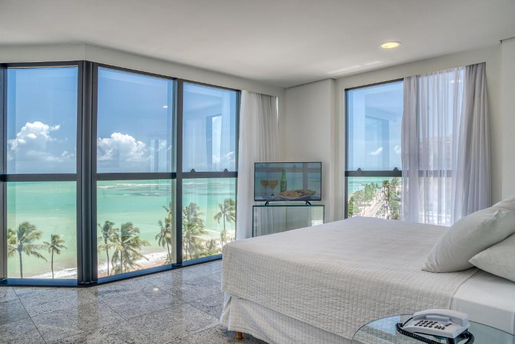 Quarto no Hotel Brisa Tower na Praia de Jatiúce, de esquina com janelas grandes que mostram a praia, as palmeiras e a vista para o mar, com uma cama e um aquário, para representar onde ficar em Maceió.