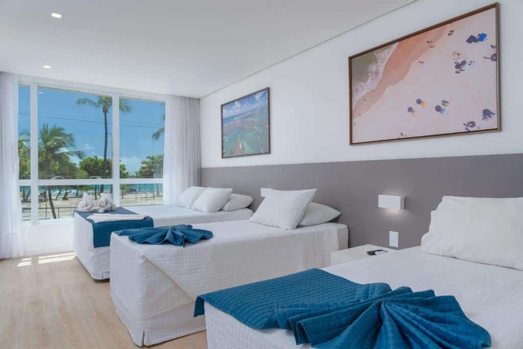 Um quarto no Hotel Praia Bonita Jangadeiros na Praia de Pajuçara, bem iluminado com três camas de solteiro, cobertores azuis, janelas grandes e arte na parede com tema de praia, para representar onde ficar em Maceió.