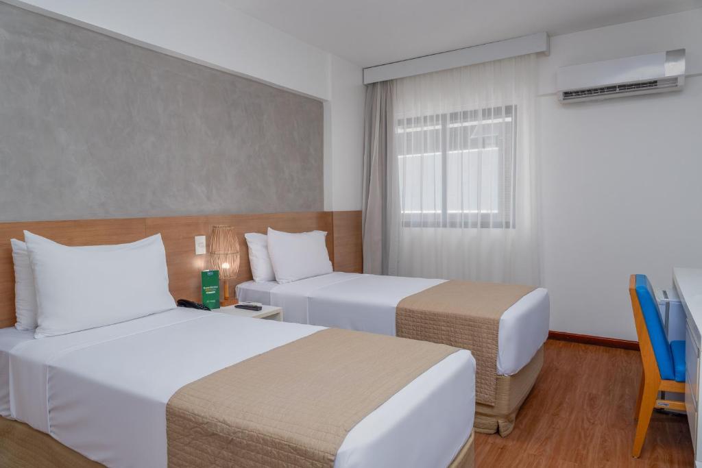 Duas camas de solteiro com roupa de cama bege em um quarto, com janela, escrivaninha, cadeira e ar-condicionado, no Palms Ponta Verde by Tropicalis na Praia de Ponta Verde, para representar onde ficar em Maceió.