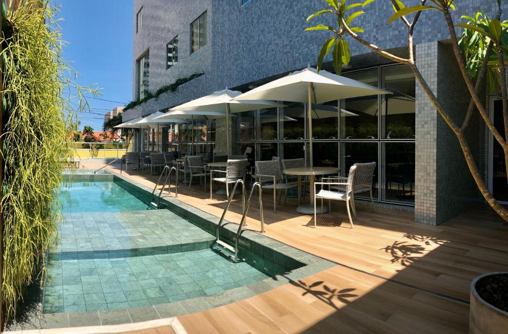 Piscina externa ao lado de um edifício moderno com mesas, cadeiras e guarda-sóis em um deck de madeira sob um céu claro, na propriedade do Flix Hotel na Praia de Jatiúca.