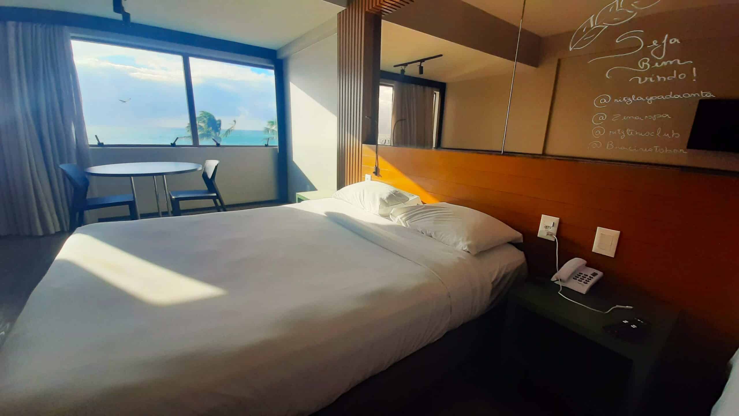 Quarto do Ritz Lagoa da Anta Hotel na Cruz das Almas, com uma cama de casal, um espelho acima da cama (como se fosse uma cabeceira) e, à esquerda, tem uma mesa com duas cadeiras e uma janela de vidro ocupando metade da parede de fora a fora com vista do mar, para representar onde ficar em Maceió.