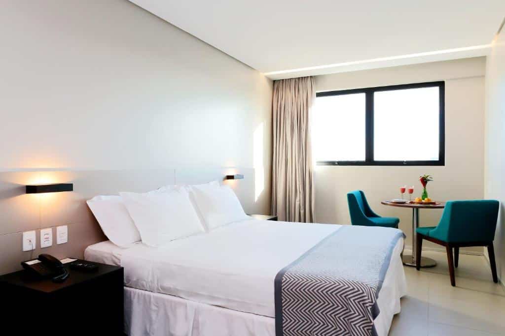 Um quarto no Porto Kaeté Hotel na Praia de Jatiúca, com uma cama bem arrumada, uma mesa de cabeceira, duas cadeiras azul-petróleo, uma mesa redonda e uma grande janela com cortinas, para representar onde ficar em Maceió.