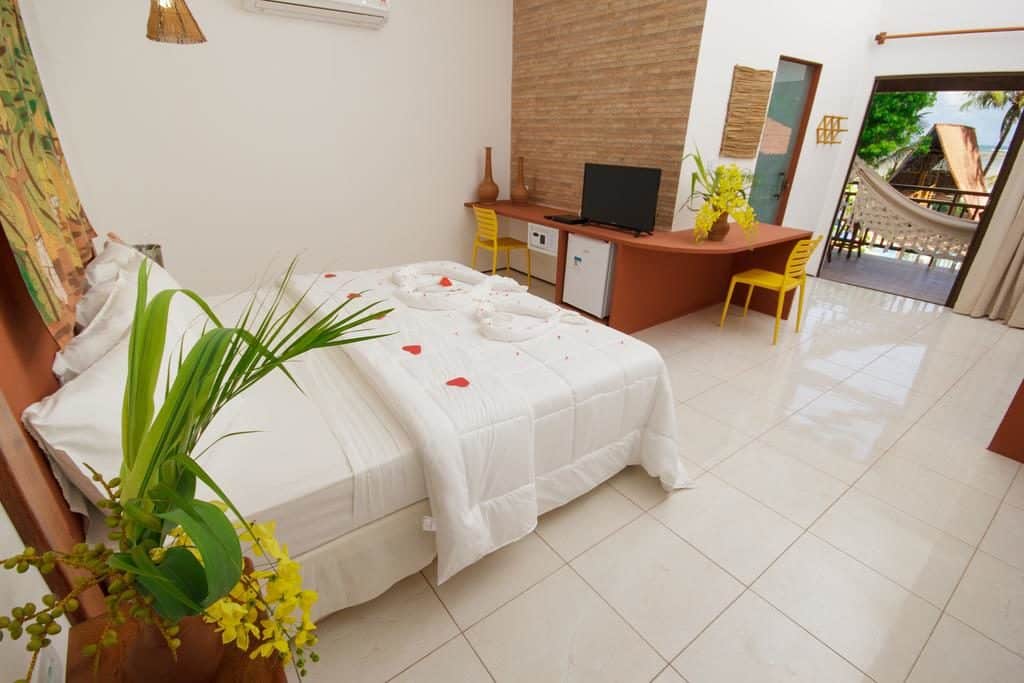 Um quarto de hotel arrumado com uma cama de casal, escrivaninha, TV, cadeiras amarelas e uma rede em uma varanda visível através de portas abertas.