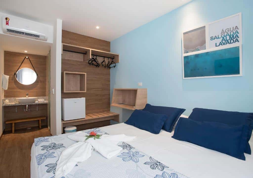 Um quarto de hotel arrumado com uma cama de casal, detalhes em azul, armário aberto, arte na parede e um banheiro adjacente com um espelho redondo.