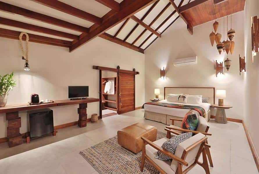 Quarto espaçoso com vigas expostas, uma cama, escrivaninha, TV, frigobar, poltrona e decoração neutra e aconchegante. Esta é uma das opções onde ficar em Maragogi.