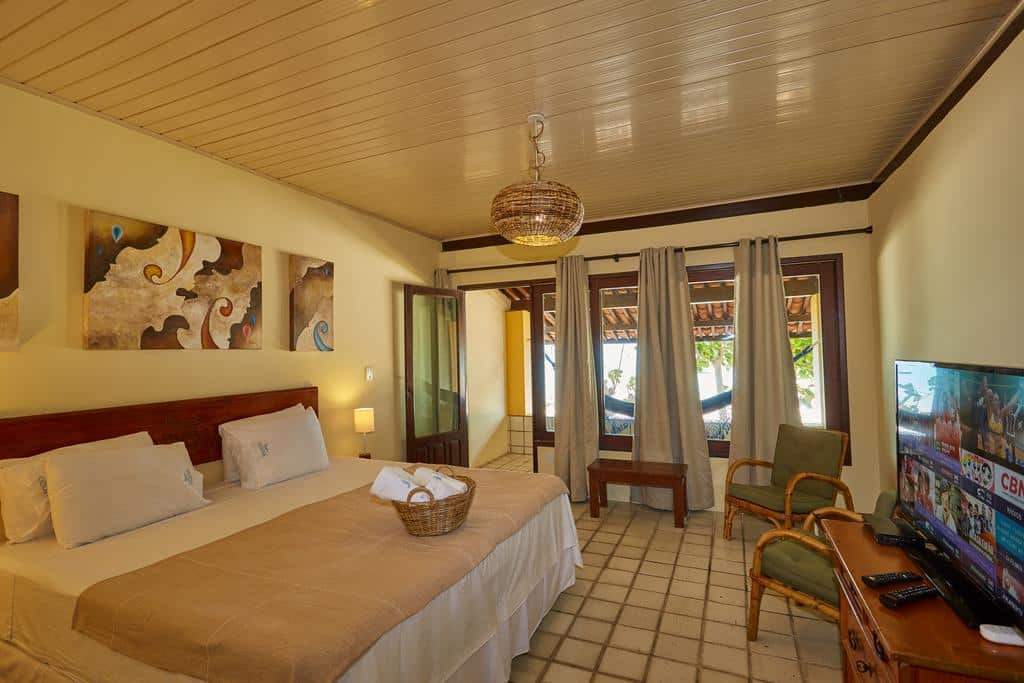 Quarto de hotel com uma cama de casal, poltrona, TV, luz de teto, arte na parede e portas de vidro que dão para um pátio. Esta é uma das opções onde ficar em Maragogi.