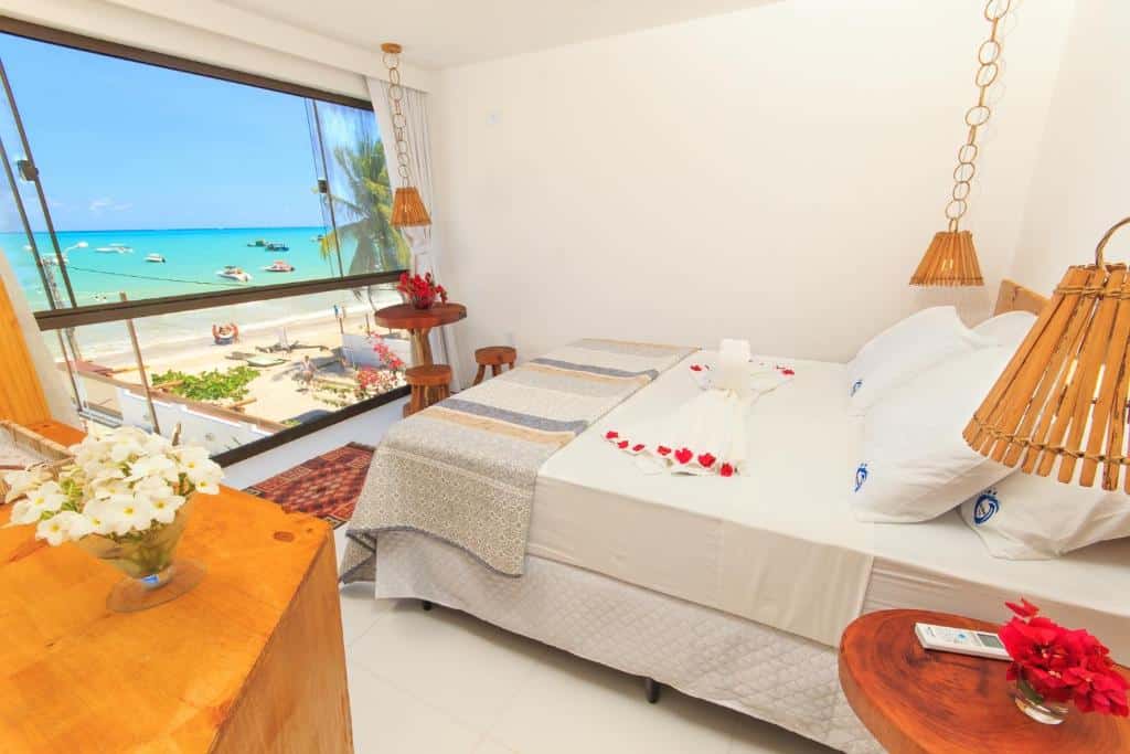 Um quarto bem iluminado com uma grande janela com vista para a praia; a cama é decorada com toalhas e pétalas de flores vermelhas. Esta é uma das opções onde ficar em Maragogi.