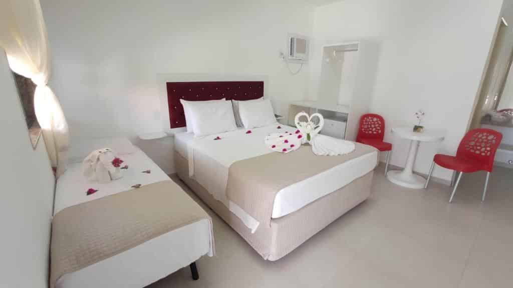 Quarto de hotel com uma cama de casal, uma cama de solteiro, cadeiras vermelhas em uma pequena mesa e cisnes de toalha nas camas.