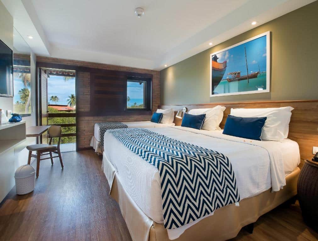 Quarto de hotel com duas camas grandes, cobertores chevron, escrivaninha, janela, porta da varanda e um quadro de barco na parede. Esta é uma das opções onde ficar em Maragogi.