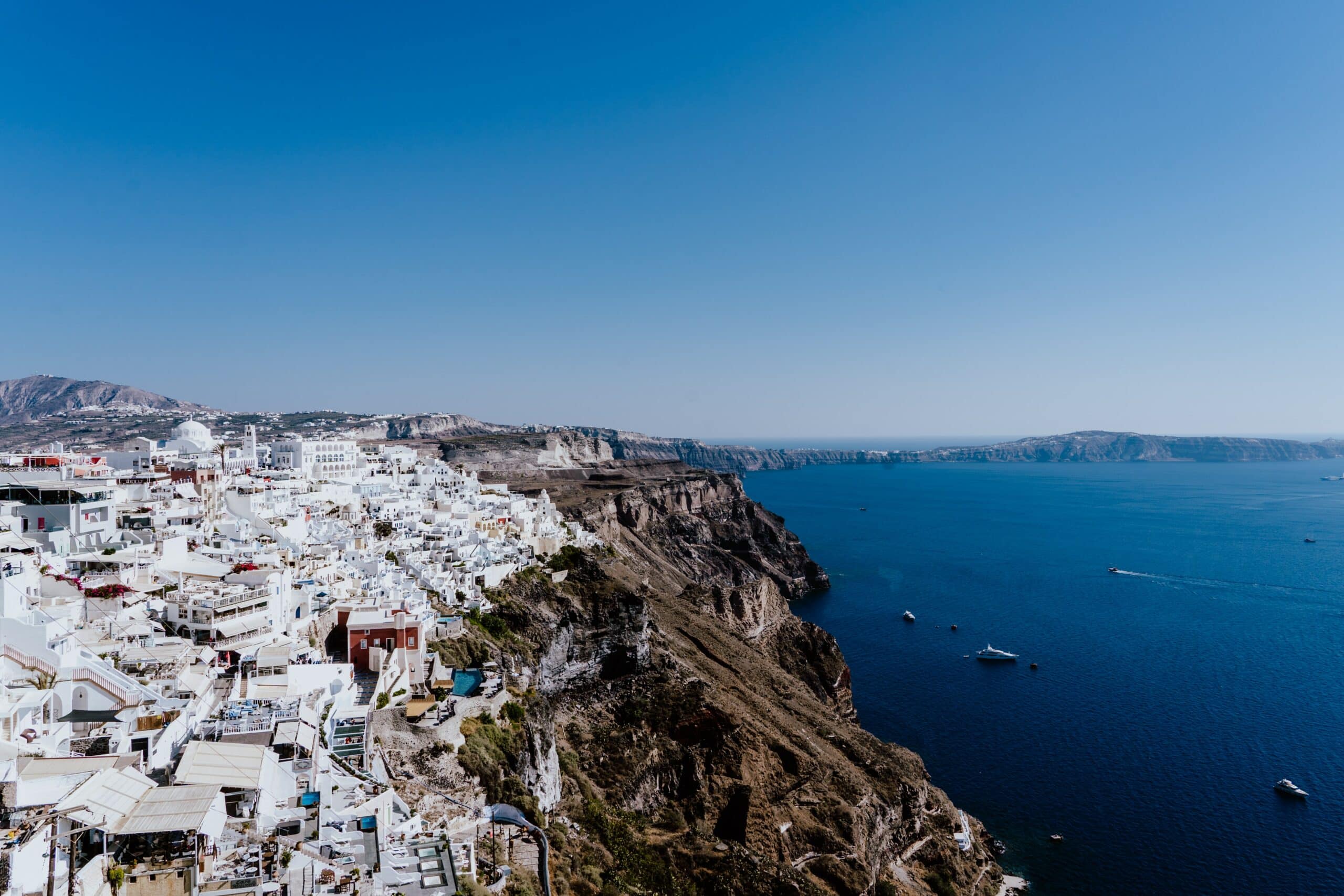 Edifícios brancos situados em um penhasco acima do mar azul com barcos, sob um céu claro, provavelmente em Santorini, Grécia. - Representa onde ficar em Santorini