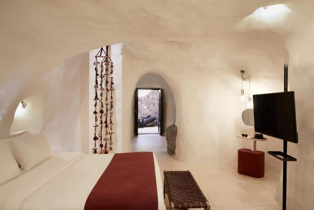 Um quarto branco em estilo de caverna com uma cama, TV, pufe e uma porta em arco que leva a outro cômodo.