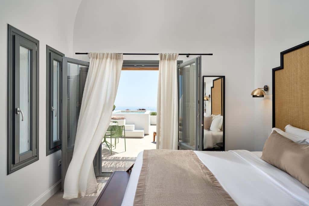 Um quarto com uma cama feita, janelas abertas, cortinas brancas e vista para um pátio com mobília externa. Hotel em onde ficar em Santorini.