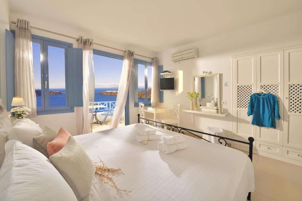 Quarto bem iluminado com uma cama feita, guarda-roupa, escrivaninha e portas da varanda que se abrem para uma vista do mar sob um céu azul.