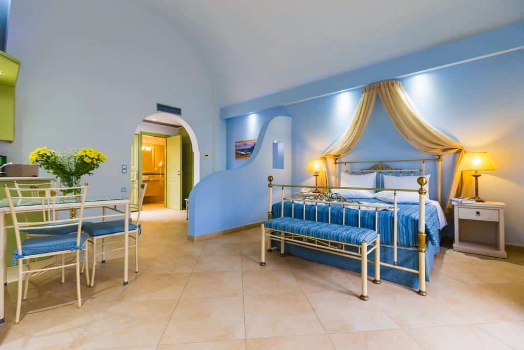 Um quarto espaçoso com uma cama azul e dourada, dossel, mesas de cabeceira, luminárias e uma pequena área de jantar com cadeiras e flores.