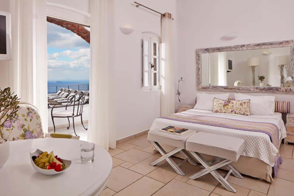 Quarto de hotel bem iluminado com cama, mesa e varanda com vista para o mar; tigela de frutas e copo de água sobre a mesa.