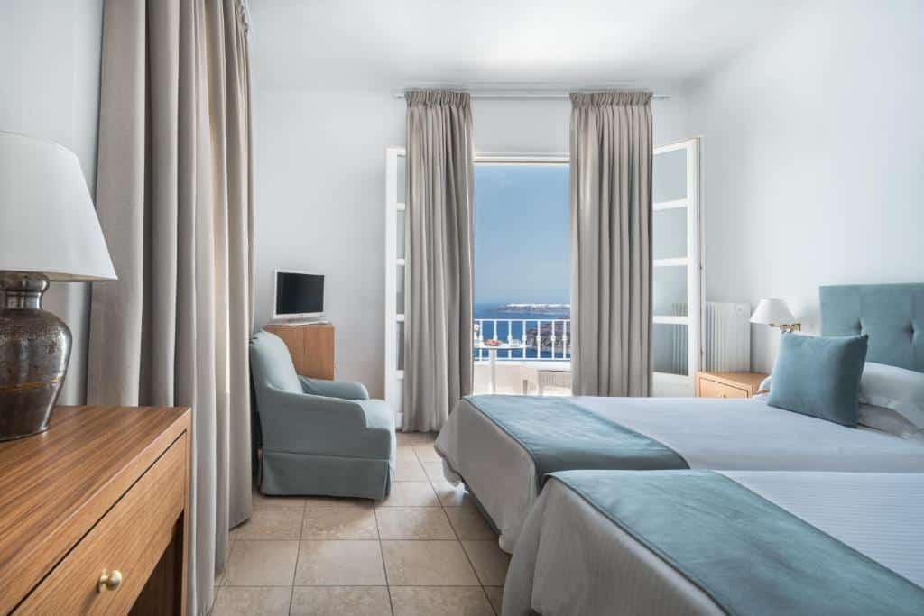 Quarto de hotel com camas de solteiro, detalhes em azul claro, poltrona, TV e portas abertas para uma varanda com vista para o mar.
