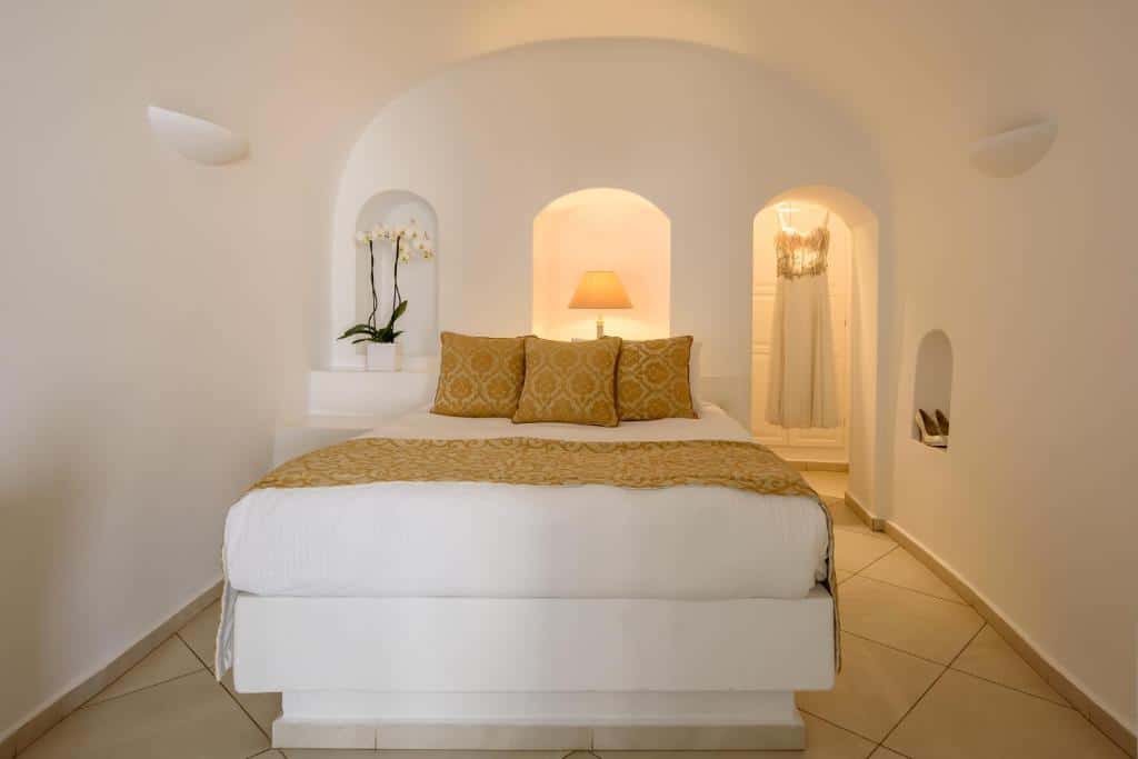 Uma cama bem arrumada com travesseiros dourados, uma orquídea branca, uma lâmpada e um vestido pendurados em uma alcova em arco.
