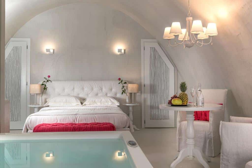 Um quarto branco com uma cama de casal, uma mesa redonda com frutas, um lustre e uma banheira de hidromassagem em primeiro plano.