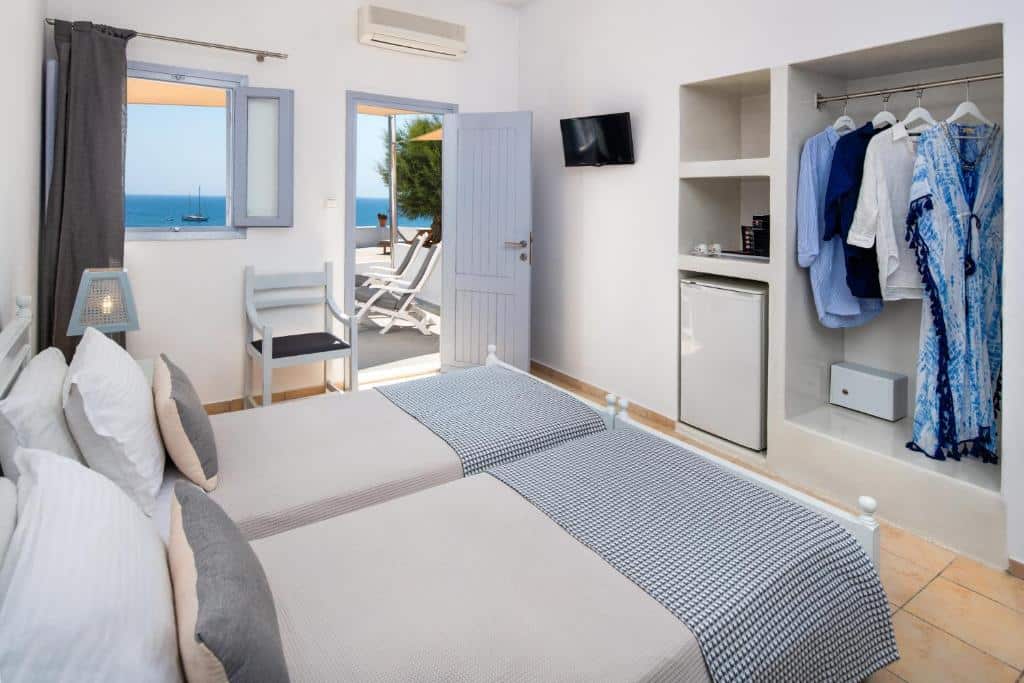 Um quarto arrumado com duas camas de solteiro, um armário aberto, um pequeno frigobar e uma porta que dá para uma varanda com vista para o mar.