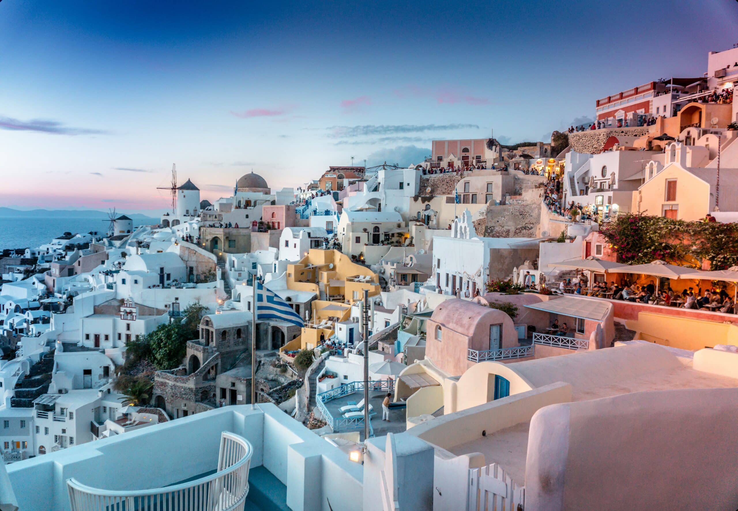 Edifícios brancos e em tons pastéis em uma encosta em Santorini, Grécia, ao pôr do sol, com o mar ao fundo. 