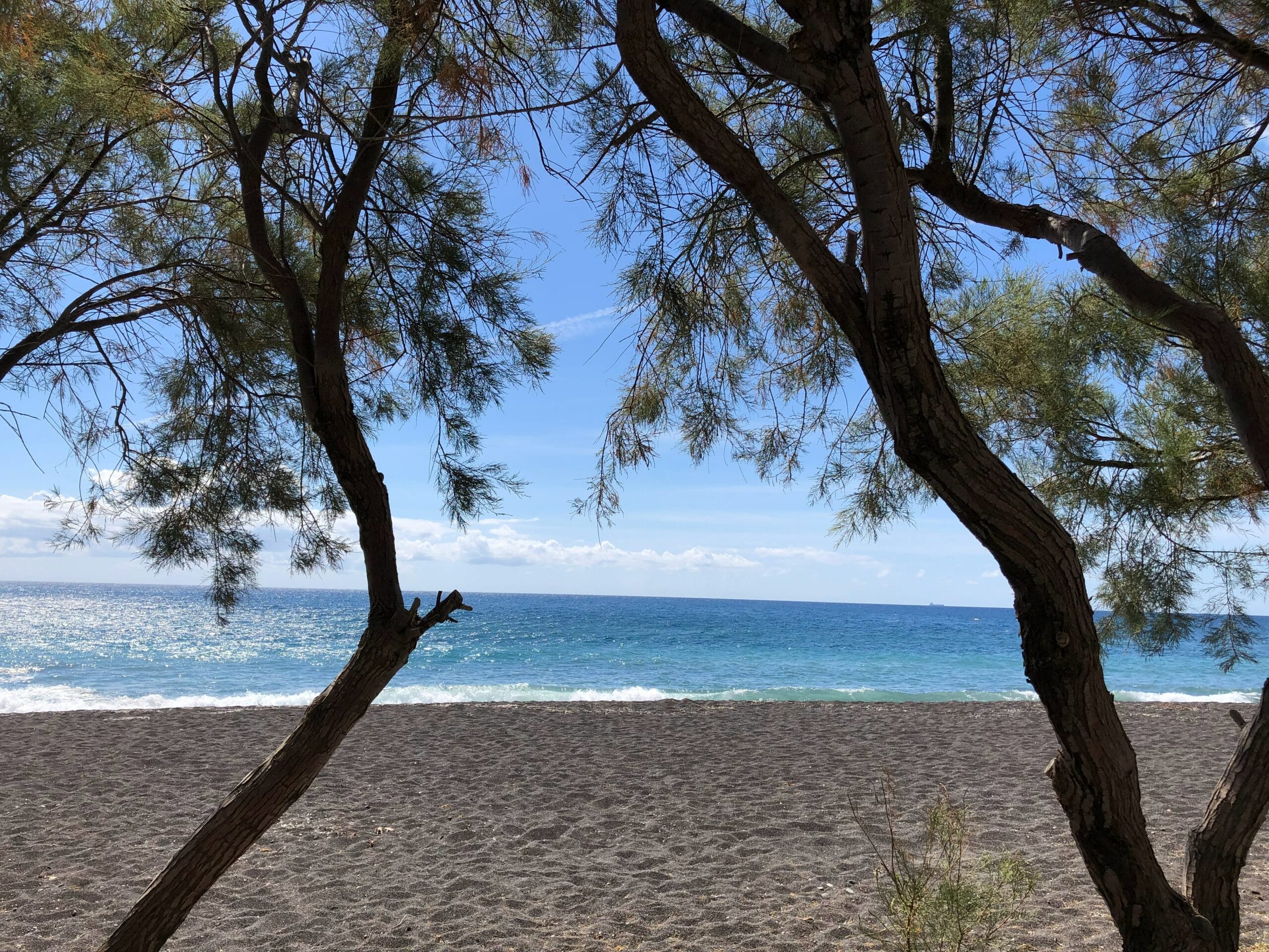 Duas árvores emolduram a vista de uma praia de areia e um oceano azul calmo sob um céu parcialmente nublado. - Representa onde ficar em Santorini
