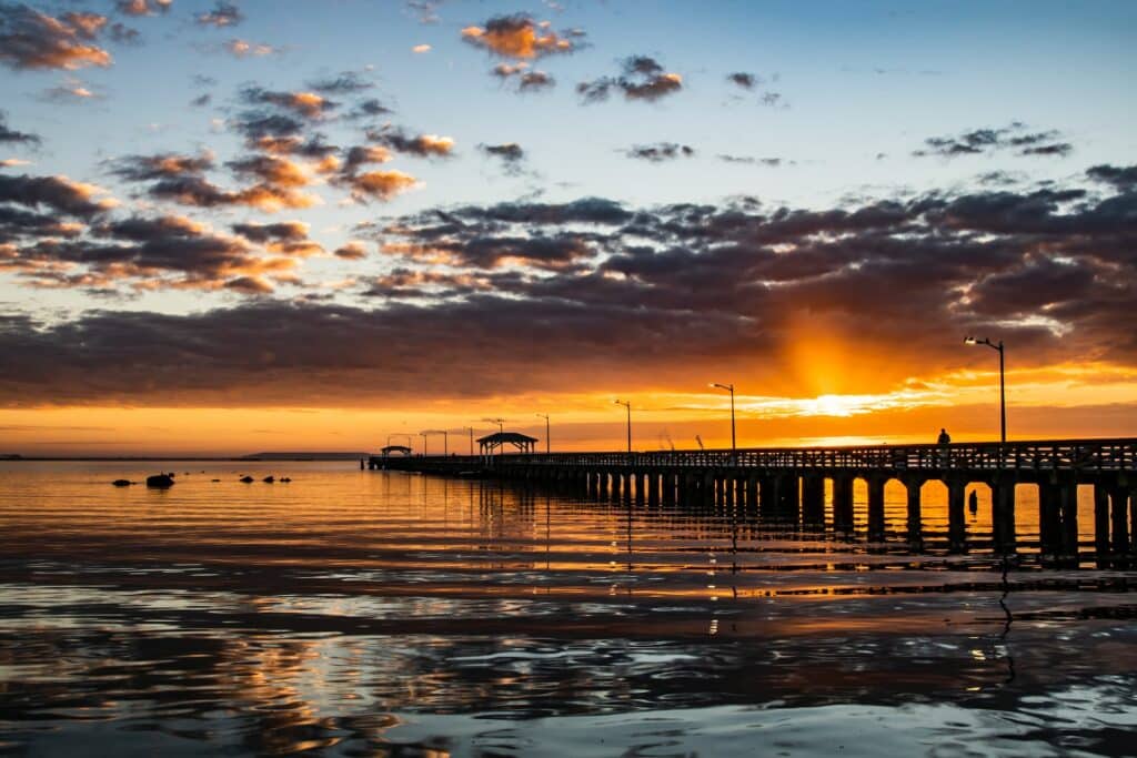 Um longo píer se estende sobre águas calmas ao pôr do sol, com nuvens dispersas e reflexos na superfície, em Ballast Point Park em South Tampa.