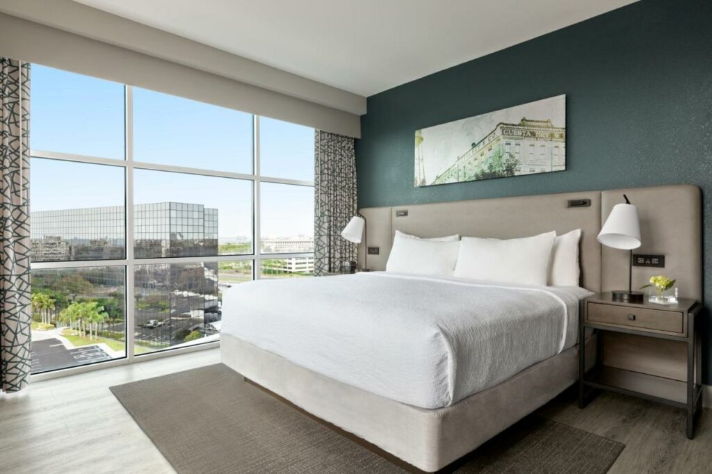 Quarto do Hotel Alba Tampa, Tapestry Collection By Hilton com uma ampla janela dando vista para a cidade, uma cama de casal, duas mesinhas de cabeceira com abajures, tudo em tons de cinza e branco, o chão é de madeira.