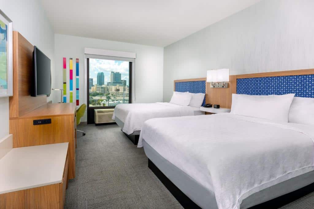 Um quarto no Hampton Inn Tampa Downtown Channel District com uma cama de casal, uma de solteiro, uma janela ampla, uma mesinha de trabalho com uma cadeira giratória, um rack e uma televisão.