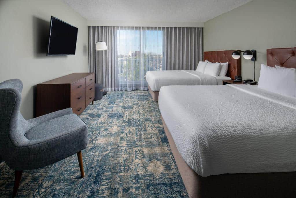 Quarto no Four Points by Sheraton Suites Tampa Airport Westshore com uma cama de casal, uma de solteiro, uma ampla sacada, uma cômoda, uma poltrona e uma televisão, tudo em tons de branco, cinza e azul.