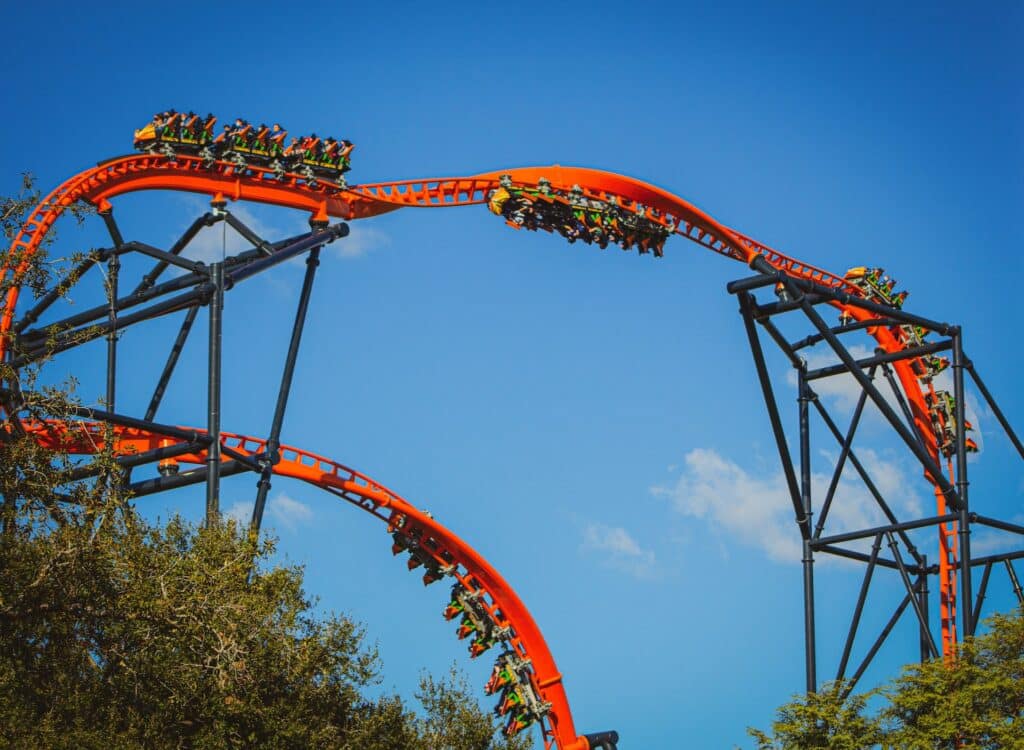 Uma montanha-russa laranja brilhante com passageiros se move ao longo de um trilho íngreme e sinuoso contra um céu azul claro e algumas árvores, no parque de diversão Busch Gardens em Tampa.